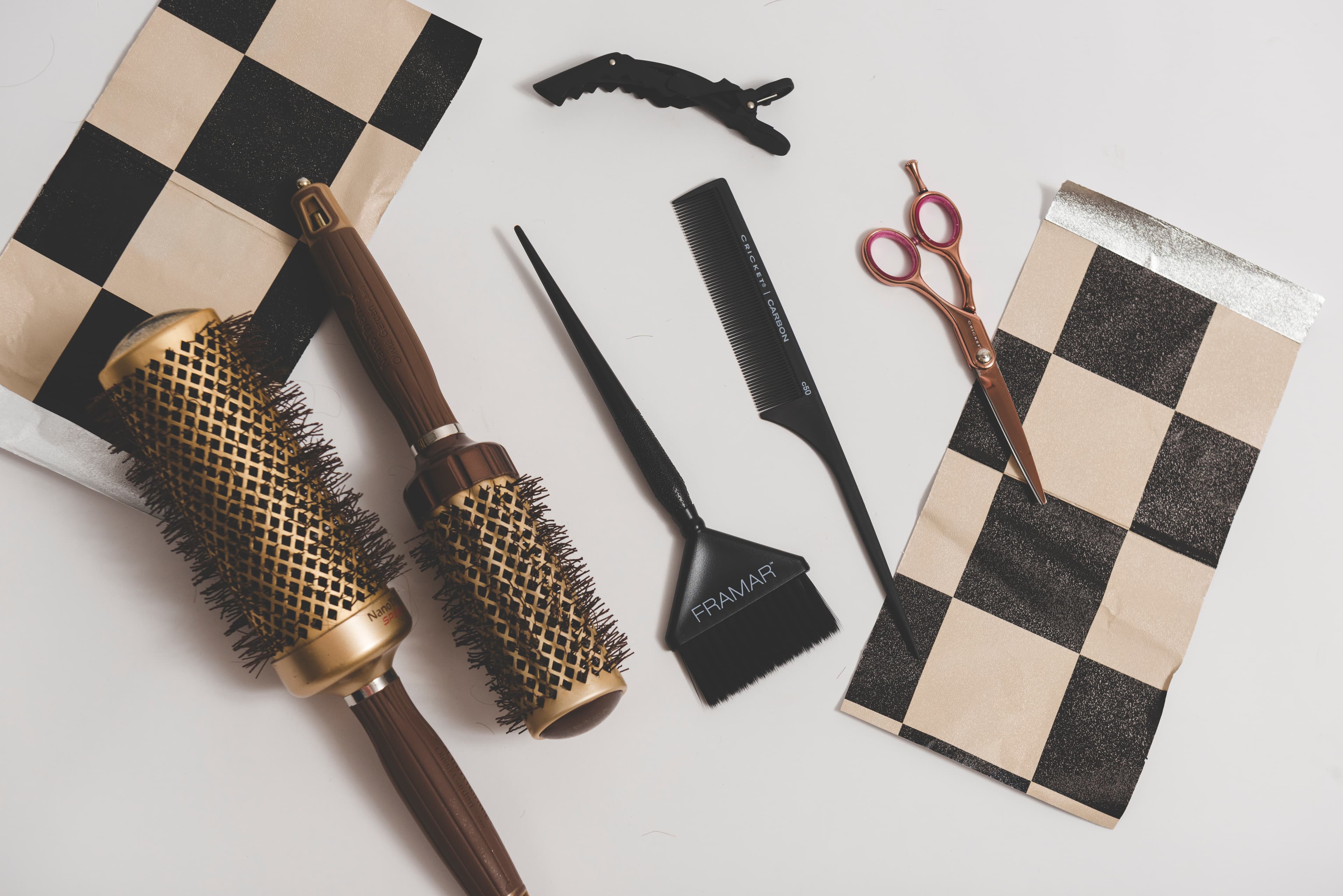 Flat-lay of professional styling brushes, shears, combs, clips, and black-and-cream checkerboard foils arranged on a work surface.